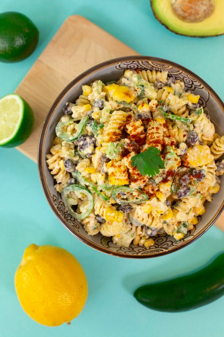An overhead shot of the Mind Over Munch recipe Easy Mexican Street Corn Pasta Salad on a blue table with sliced limes, avocado, and other veggies.