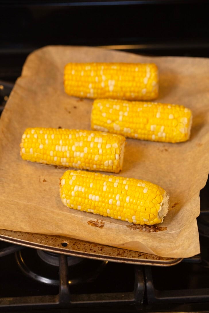 A photo of four corncobs on a baking sheet lined with parchment paper coming out of the oven. They're roasted to perfection.