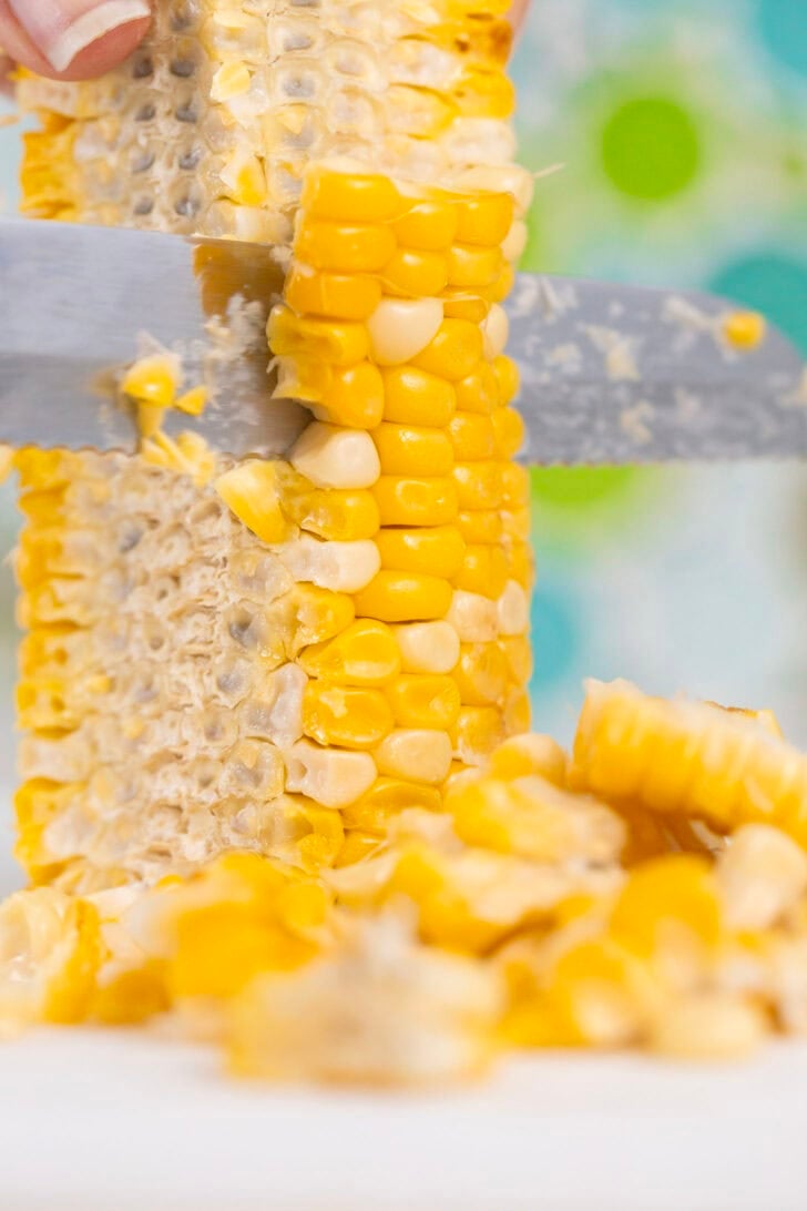 A photo of Alyssia using a small serrated knife to carefully slice the corn off of the cob so as to keep it held together in chunks rather than individual kernels. 