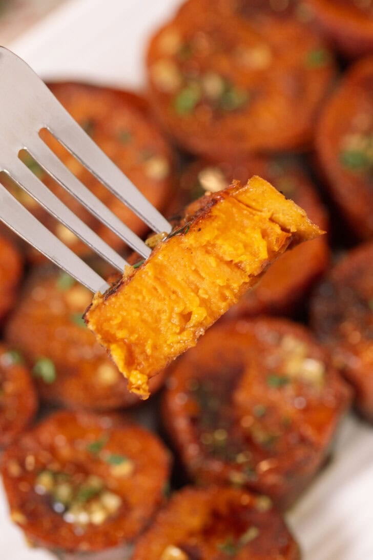 A slice of sweet potato on a fork, caramelized rounds of maple brown butter melting sweet potatoes with glaze and herbs in the background.