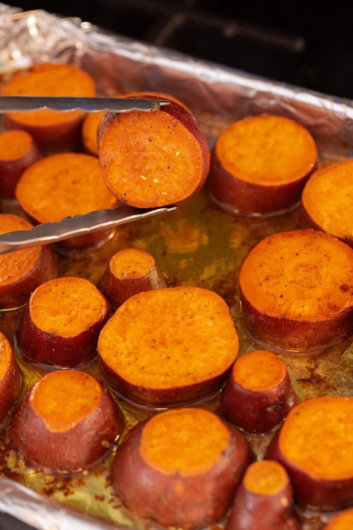 Using tongs to flip caramelized sweet potato rounds in a baking dish.