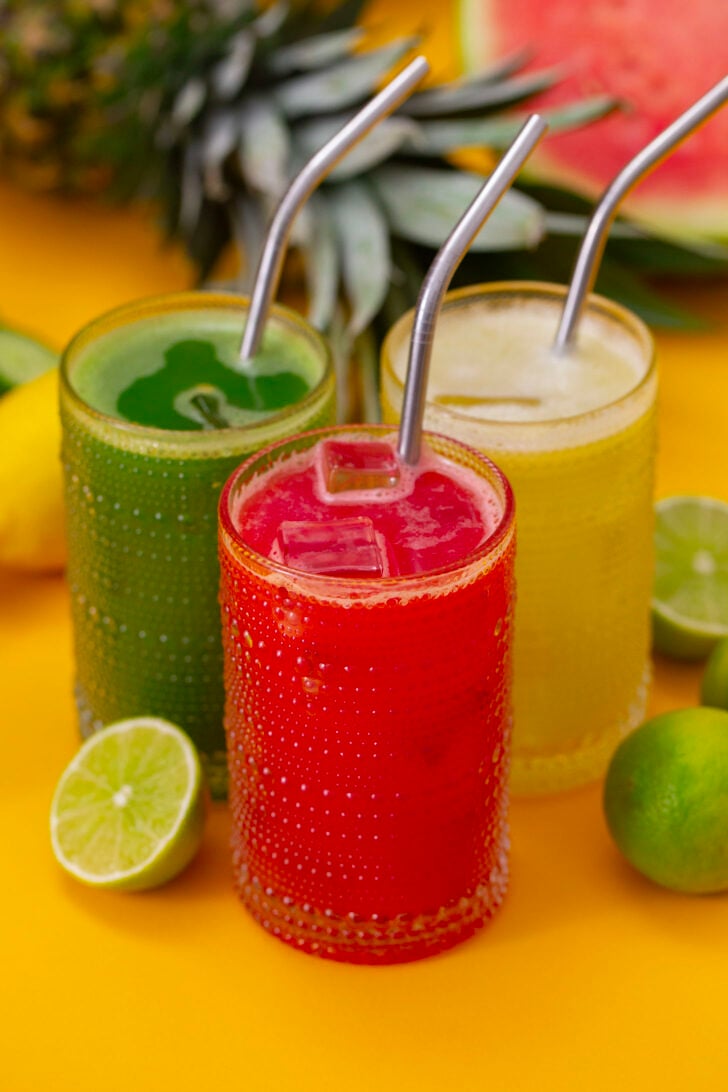 Three glasses agua fresca, on a yellow counter surrounded by ingredients.