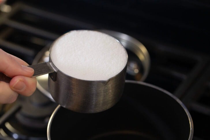 Alyssia holding a measuring cup filled with sugar over a pot on the stove.