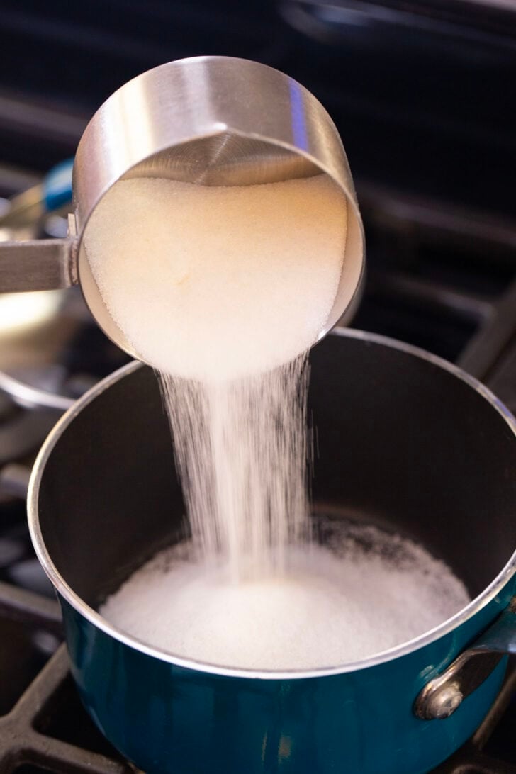 Alyssia pouring a measuring cup filled with sugar into a pot on the stove.