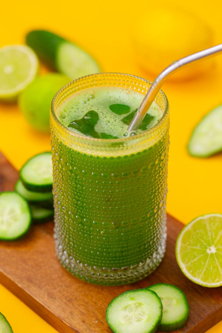 Cucumber agua fresca in a glass, sitting on a cutting board surrounded by ingredients.