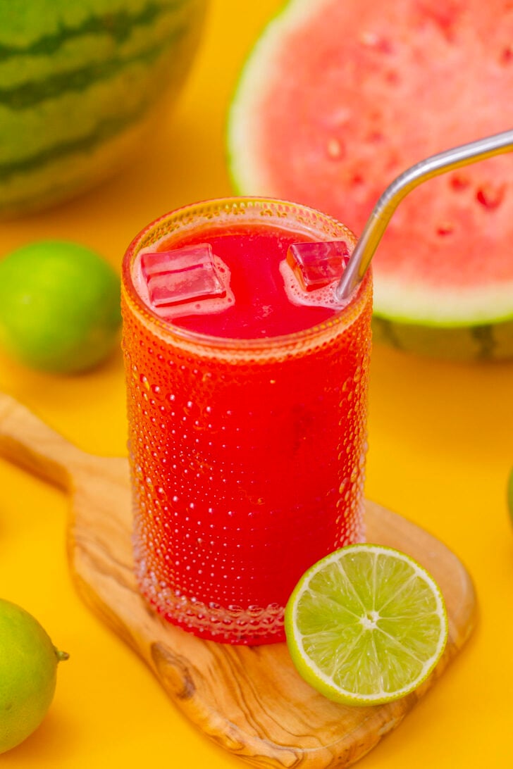 Watermelon agua fresca in a glass, sitting on a cutting board surrounded by ingredients.