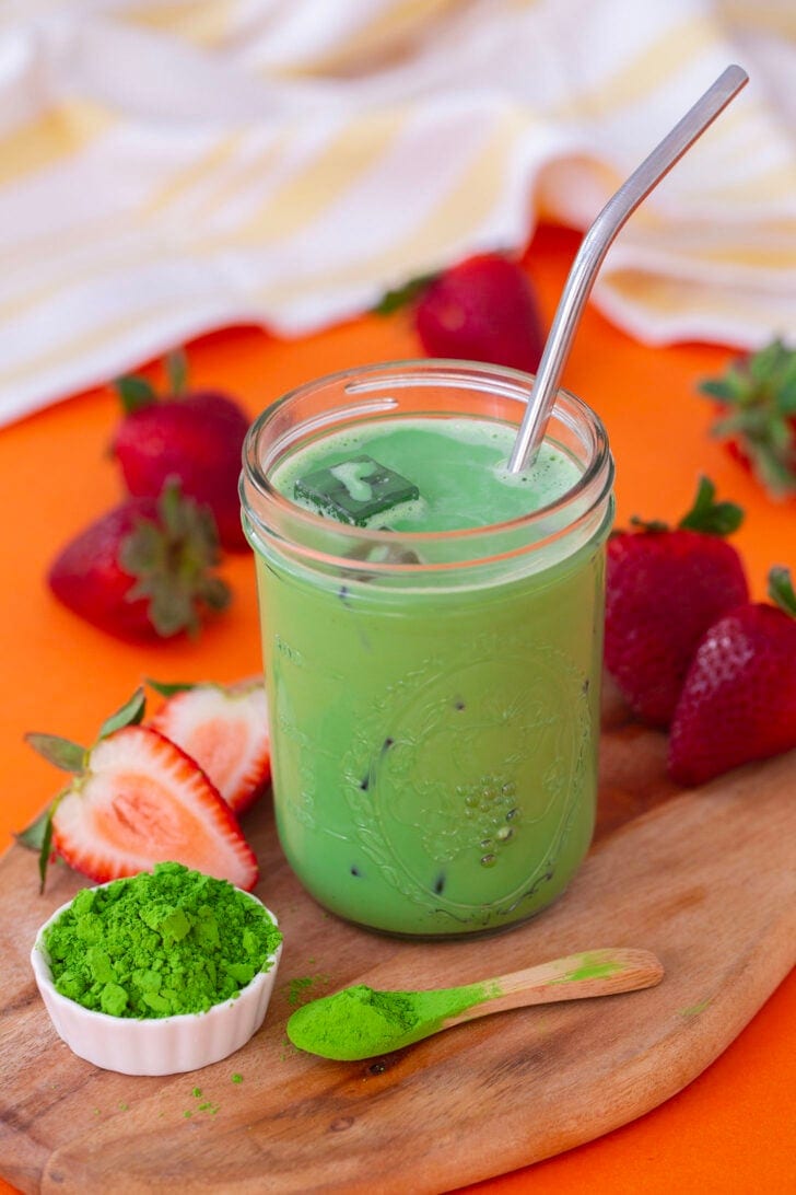 A shot of the iced matcha latte on a wooden cutting board, surrounded by powdered matcha and fresh strawberries.