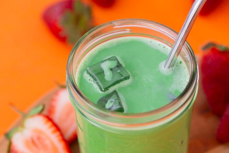 A shot of an iced matcha latte on an orange table, surrounded by fresh strawberries.