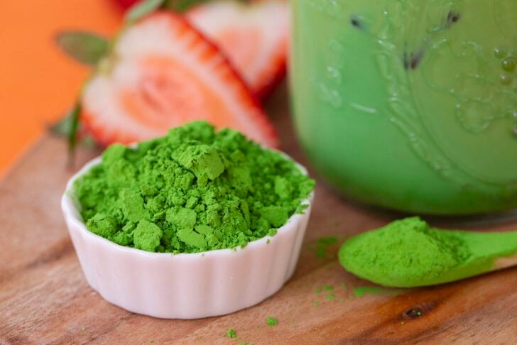 Matcha latte in a glass jar with a bowl of powdered matcha on the side, as well as a mini spoonful of matcha.