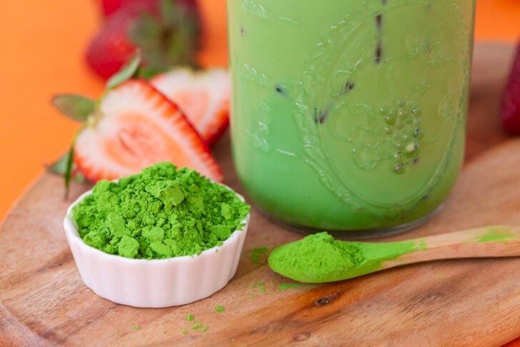 Matcha latte in a glass jar with a bowl of powdered matcha on the side, as well as a mini spoonful of matcha.
