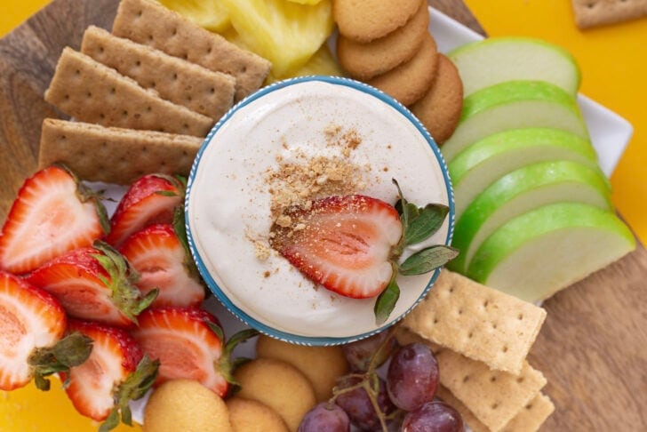 No bake cheesecake dip in a blue bowl, surrounded by dippers such as green apples, strawberries, and graham crackers and nilla wafers.