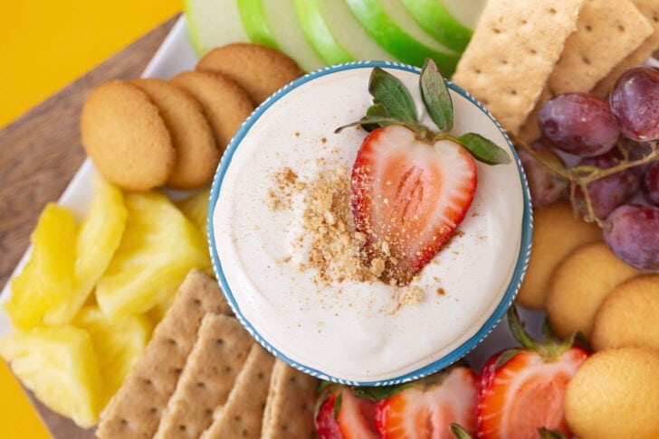 No bake cheesecake dip in a blue bowl, surrounded by dippers such as apples, strawberries, pineapple, and graham crackers.
