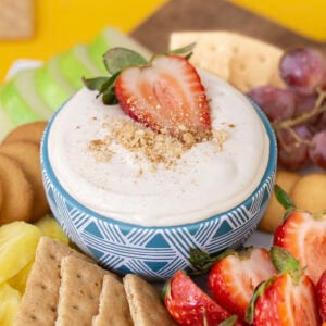 Cheesecake dip in a blue bowl, surrounded by dippers such as apples, strawberries, and graham crackers.