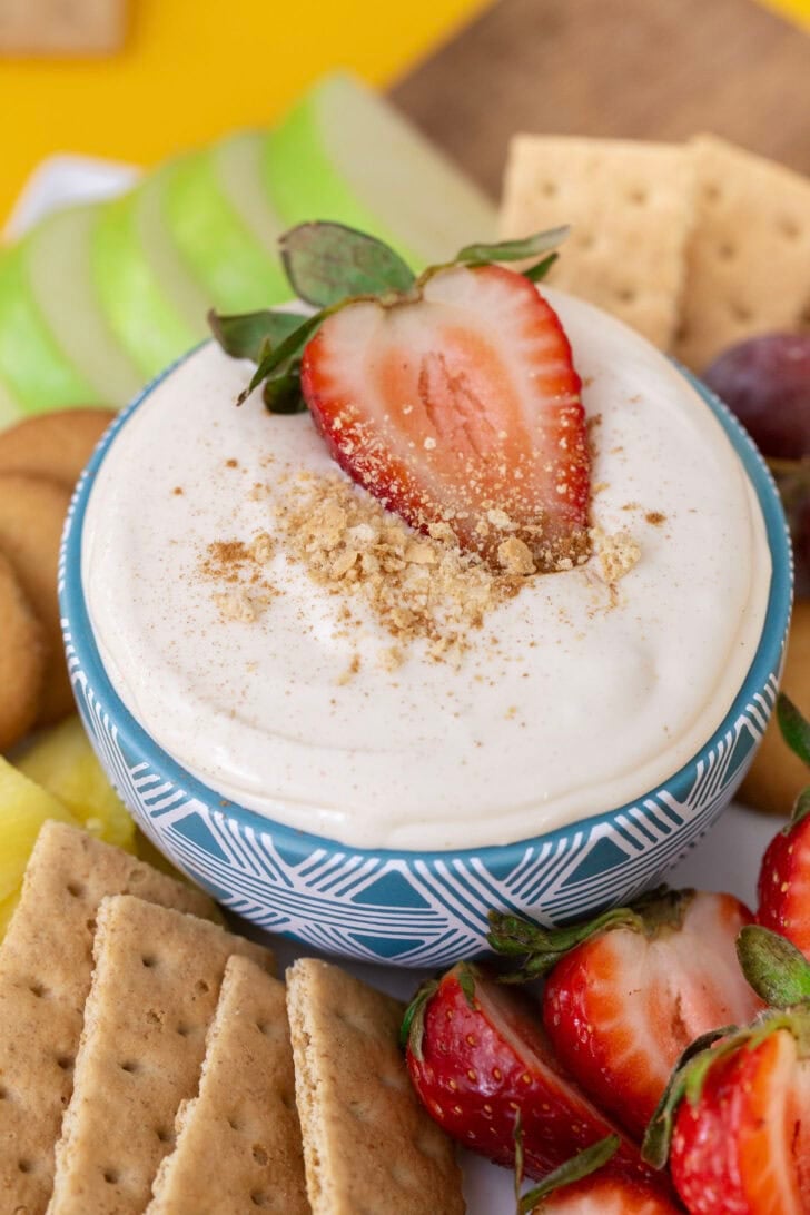 No bake cheesecake dip in a blue bowl, surrounded by dippers such as apples, strawberries, and graham crackers.