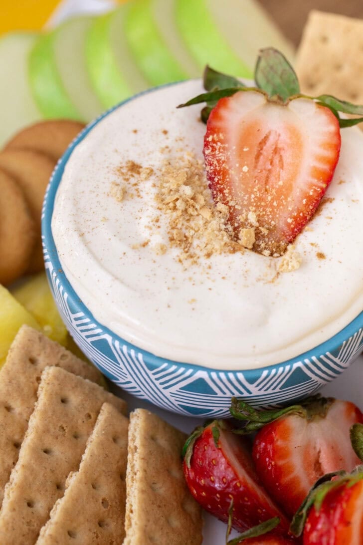 No bake cheesecake dip in a blue bowl, surrounded by dippers such as green apples, strawberries, graham crackers and nilla wafers.