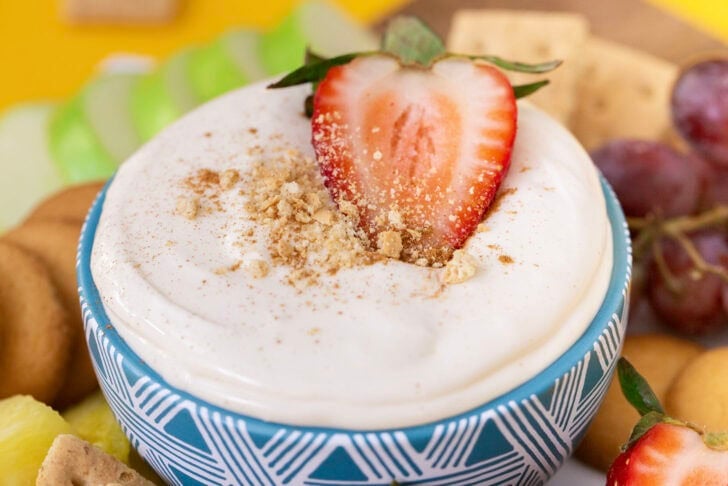 A side view of a blue bowl filled with no bake cheesecake dip, topped with a strawberry, surrounded by various dippers.