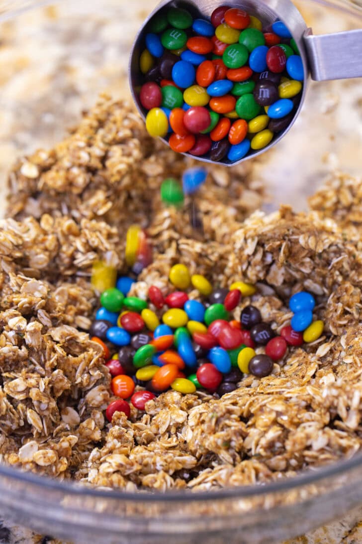 Using a measuring cup to add mini M&Ms to the protein ball oat mixture.