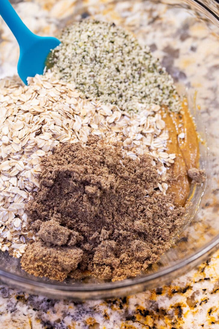 Adding rolled oats, ground flaxseed, and hemp hearts to the peanut butter mixture in a glass bowl.