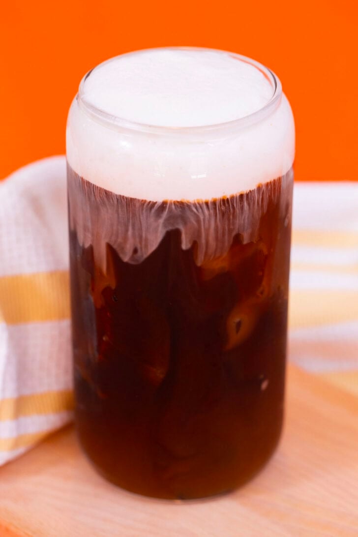 A glass of iced coffee with a vanilla cold foam cap on an orange background.
