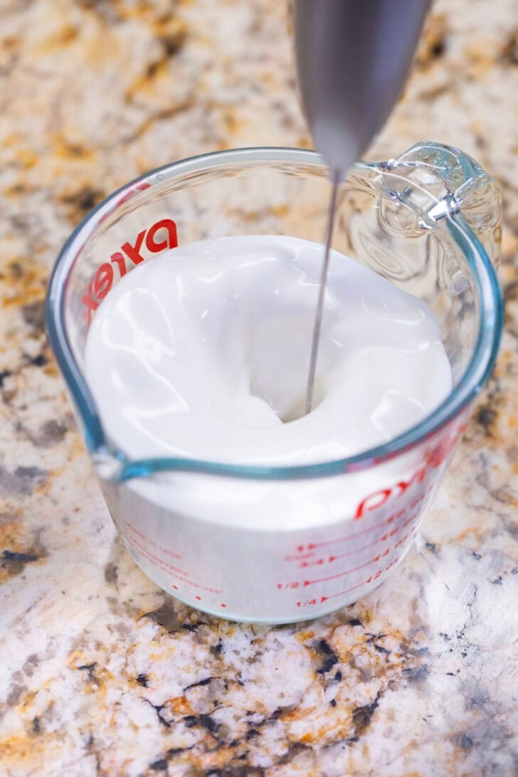 Alyssia using a hand frother to whip up the vanilla sweet cream cold foam in a glass measuring cup.