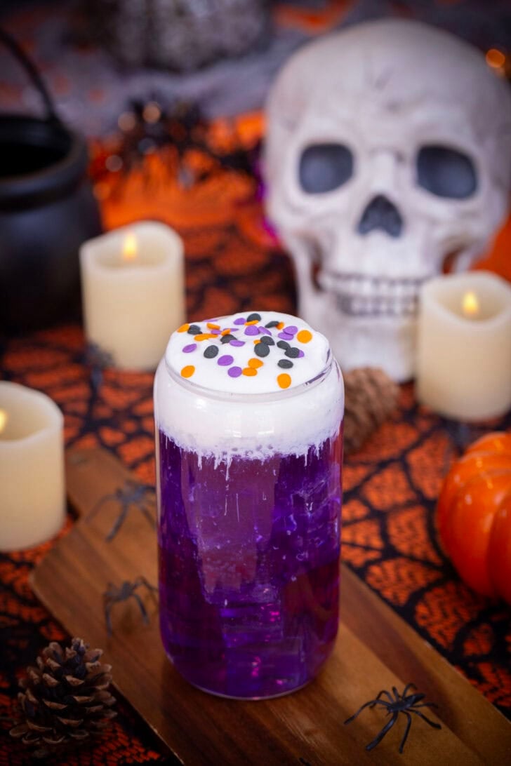 A shot of a purple halloween themed witch's brew dirty soda on a table with halloween decorations