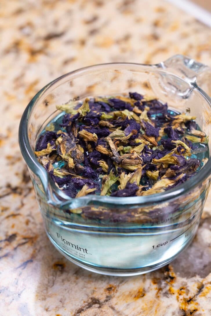 A glass measuring cup filled with hot water and steeping butterfly pea tea leaves.