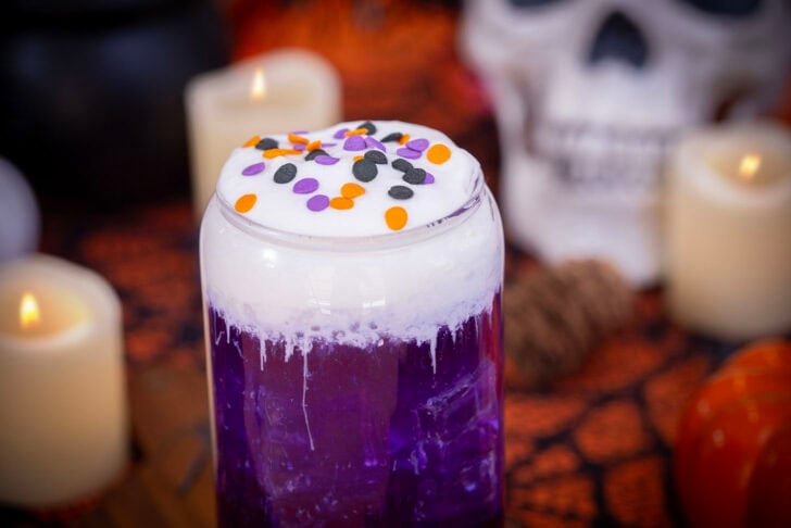 A landscape image of a purple halloween themed witch's brew dirty soda on a table with halloween decorations