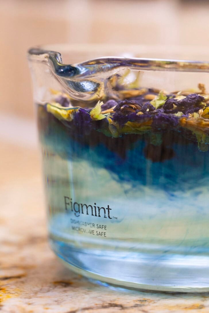 A glass measuring cup filled with hot water and steeping butterfly pea tea leaves, the blue color of the tea beginning to show.