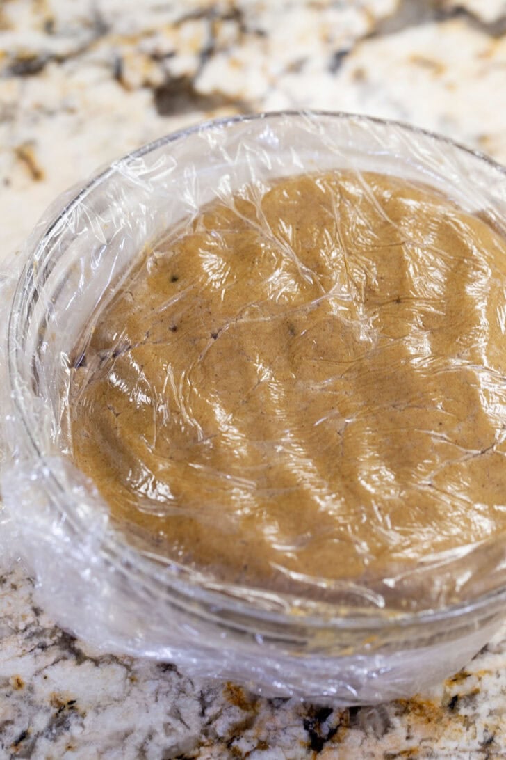 A glass bowl of eggless gingerbread cookie dough covered tightly with plastic wrap, ready to chill in the fridge.