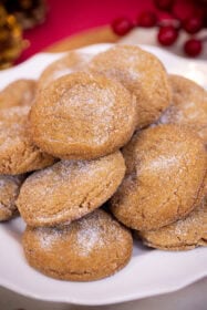 A stack of sugar-coated gingerbread cookies with crackled tops on a white plate, with red berries in the background.