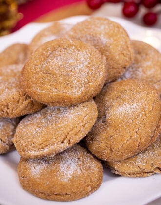 A stack of sugar-coated gingerbread cookies with crackled tops on a white plate, with red berries in the background.