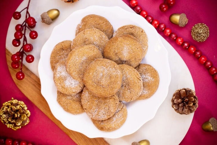An overhead shot of eggless gingerbread cookies on a white plate, surrounded by pinecones, red berry garland, and gold acorns.