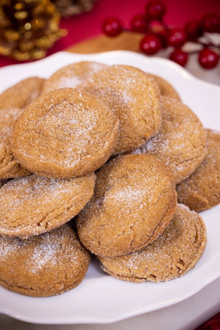 Soft, sugar-coated gingerbread cookies stacked on a white plate in front of a red holiday backdrop.
