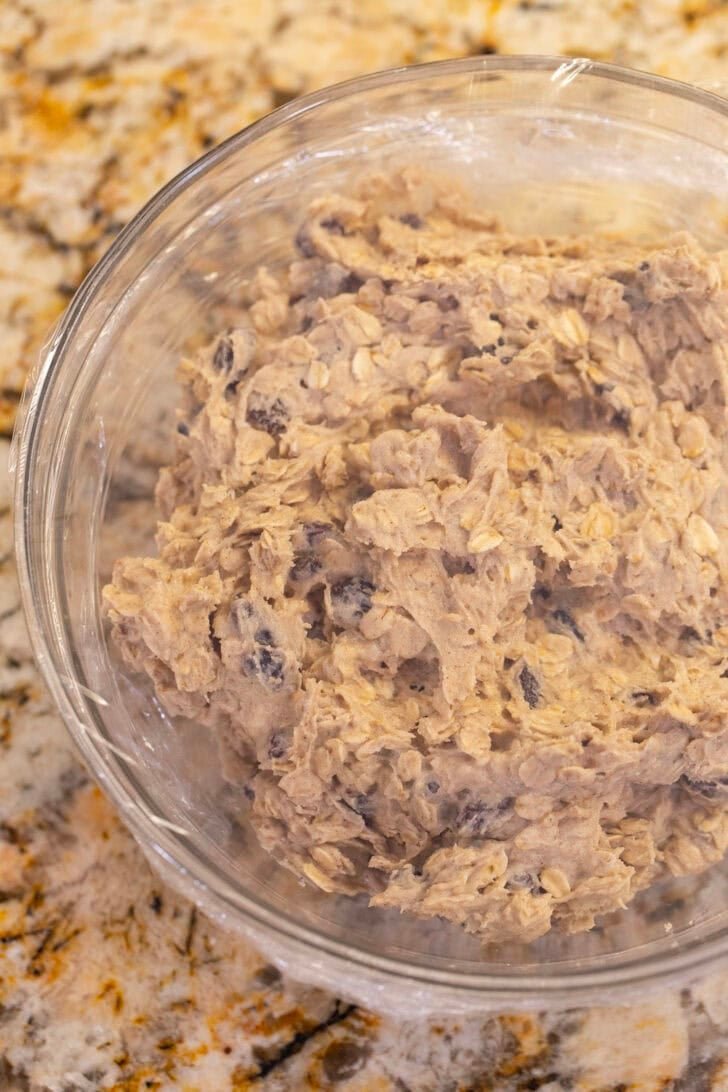 A glass bowl of oatmeal raisin cookie dough covered with plastic wrap, ready to chill.