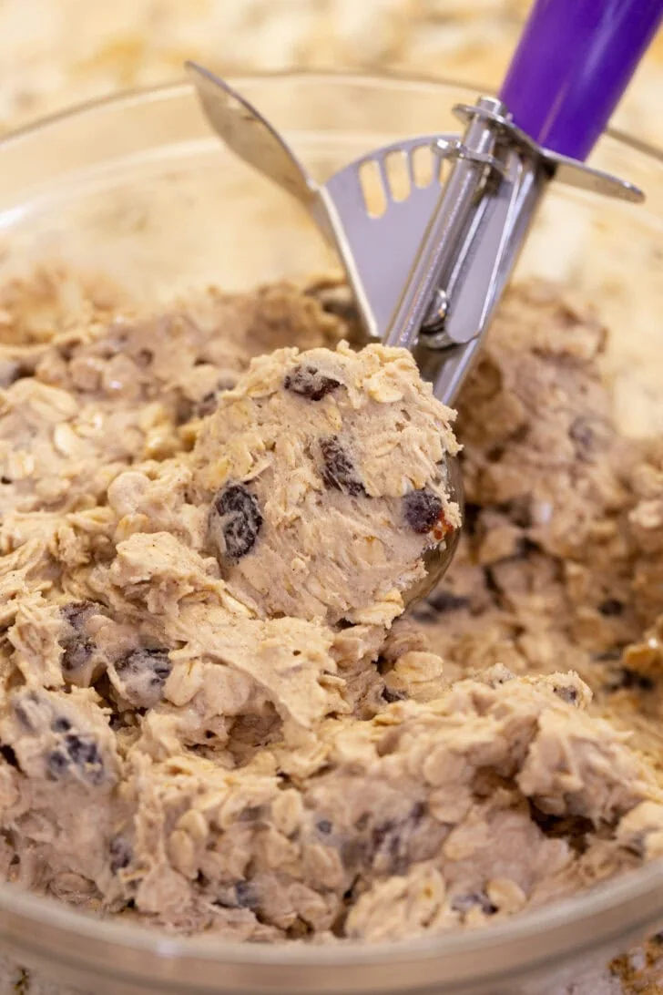 A cookie scoop filled with oatmeal raisin cookie dough over a glass bowl.