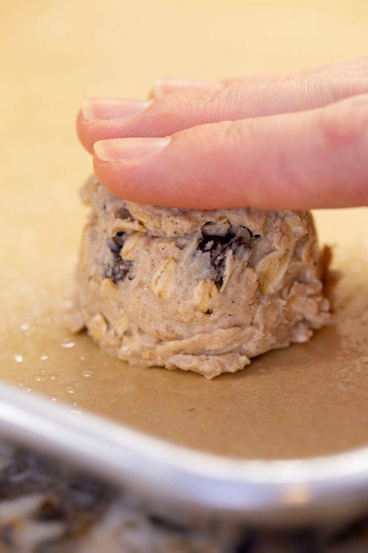 Pressing a scoop of oatmeal raisin cookie dough into a thick disc on a parchment-lined baking sheet.