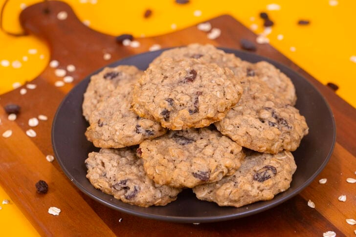 A dark plate of eggless oatmeal raisin cookies on a wooden board with oats and raisins on a yellow backdrop.