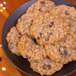 A plate of soft and chewy eggless oatmeal raisin cookies on a wooden board with oats and raisins on a yellow background.