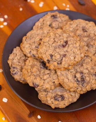 A plate of soft and chewy eggless oatmeal raisin cookies on a wooden board with oats and raisins on a yellow background.