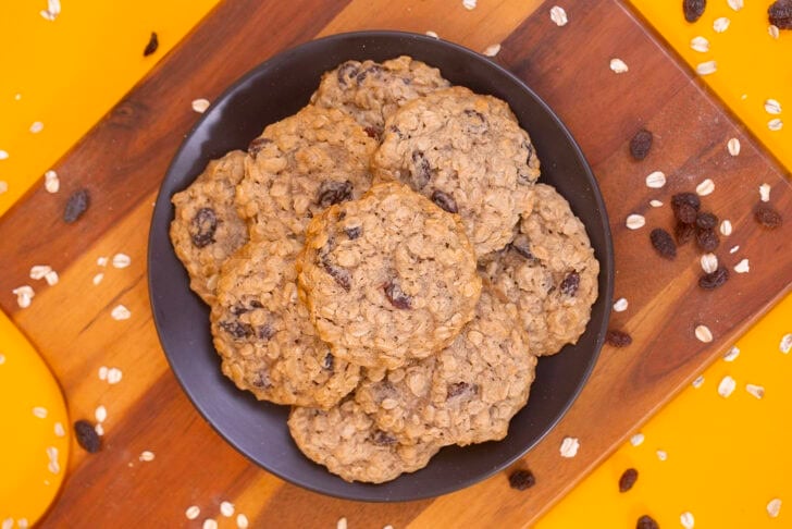 An overhead shot of a plate of oatmeal raisin cookies on a wooden board surrounded by scattered oats and raisins.