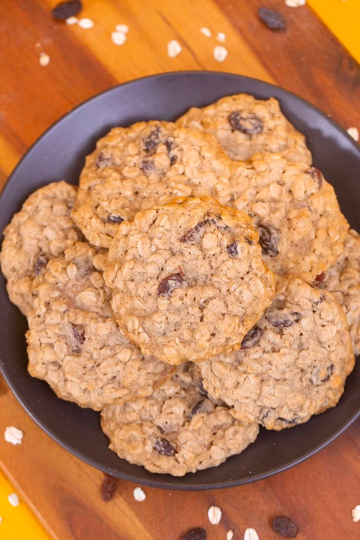 An overhead shot of chewy oatmeal raisin cookies stacked on a dark plate on a wooden cutting board.