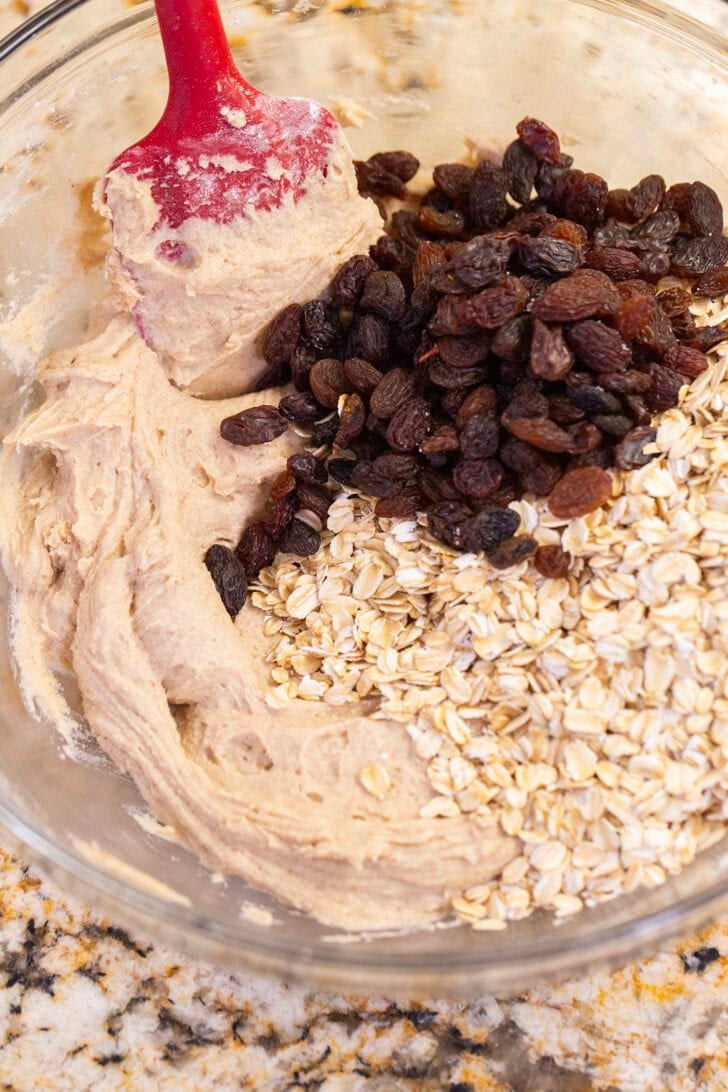 Old-fashioned rolled oats and raisins added to eggless cookie dough in a glass bowl.