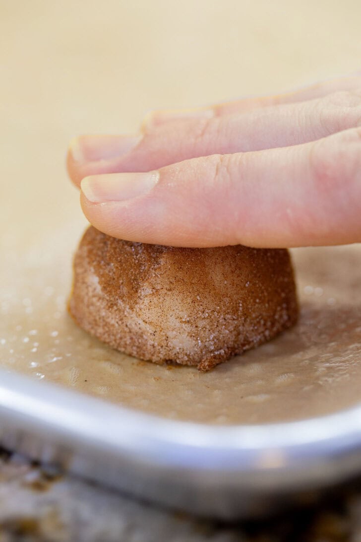 Pressing a cinnamon-sugar-coated dough ball into a thick disc on a parchment-lined baking sheet.