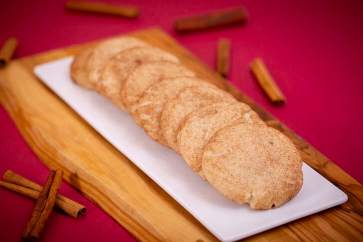 A row of eggless snickerdoodles on a white platter and wooden board, with cinnamon sticks on a red backdrop.