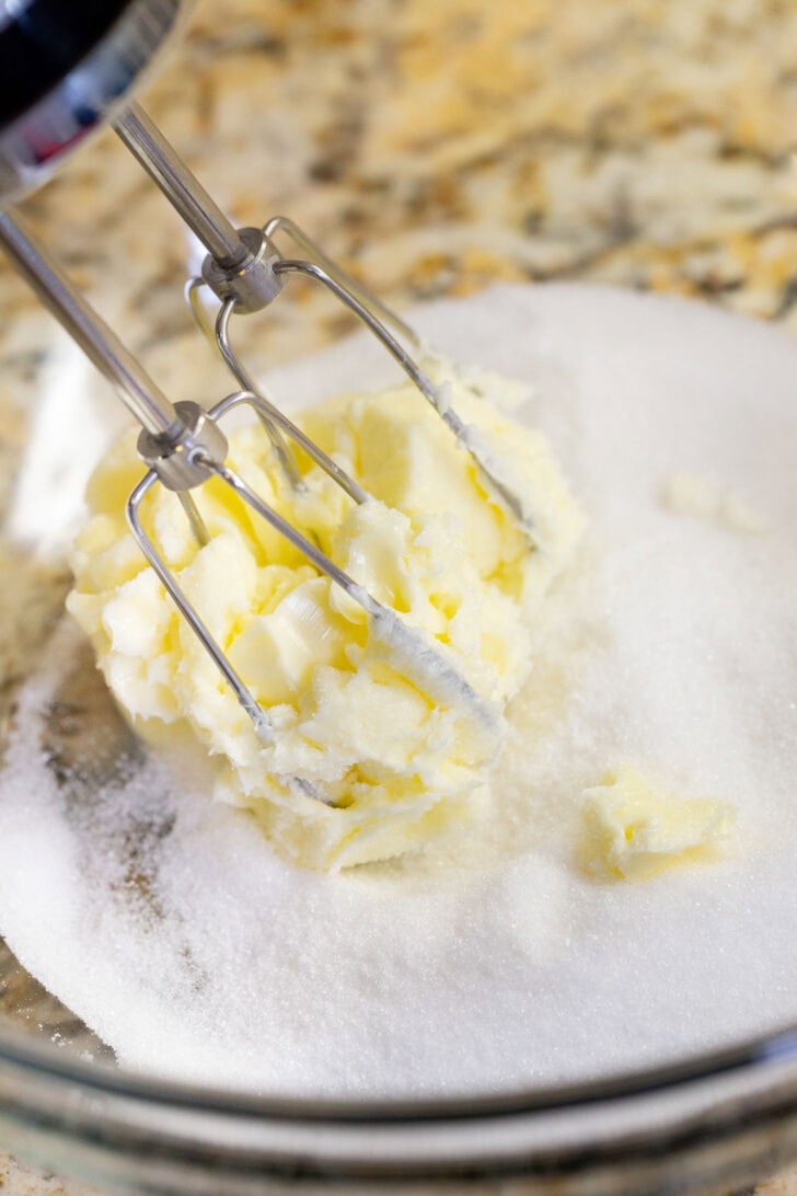 A hand mixer beating softened butter and granulated sugar together in a glass bowl.