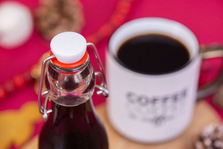 A high-angle shot of a glass bottle filled with gingerbread simple syrup next to a white coffee mug and gingerbread cookies.