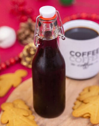 A side shot of homemade gingerbread simple syrup in a glass bottle, surrounded by gingerbread cookies with a cup of coffee behind it.