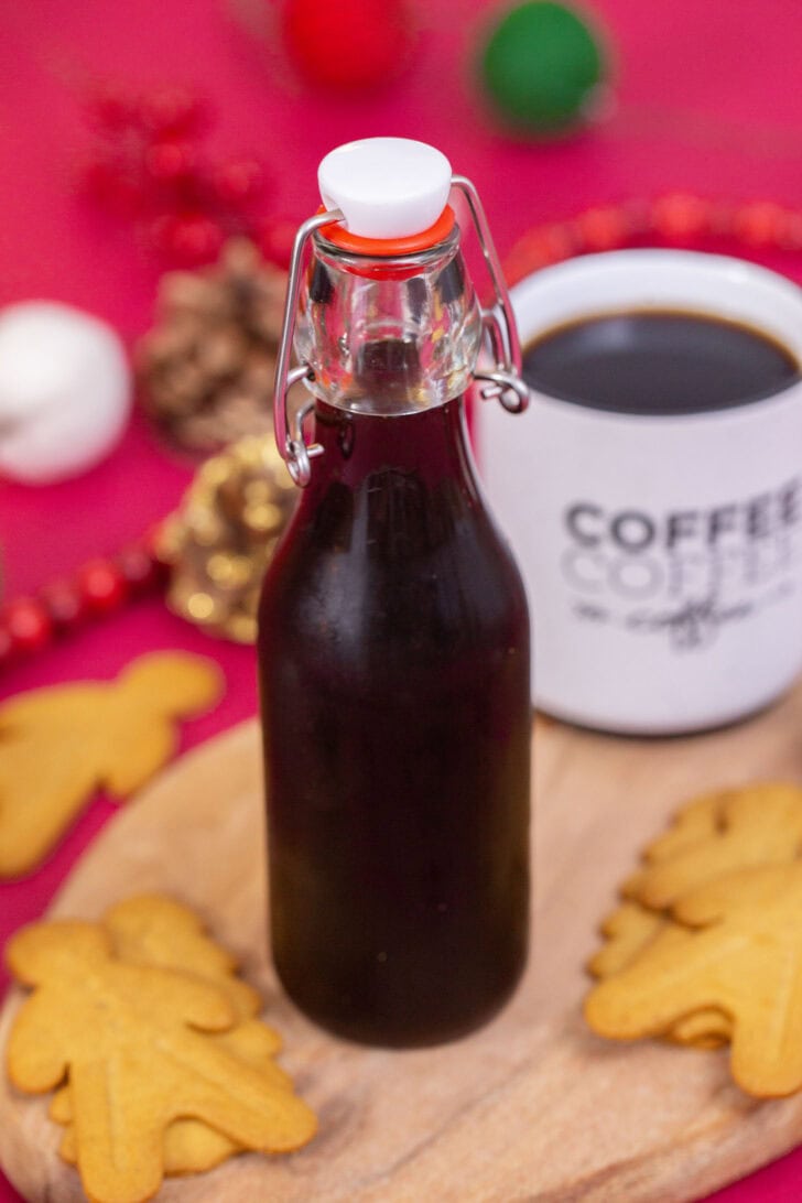 A side shot of homemade gingerbread simple syrup in a glass bottle, surrounded by gingerbread cookies with a cup of coffee behind it.