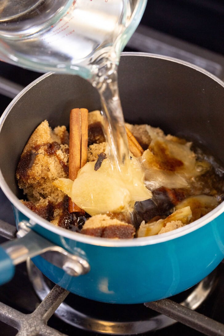 Pouring water from a glass measuring cup into a saucepan of brown sugar, molasses, ginger, and whole spices for gingerbread simple syrup.