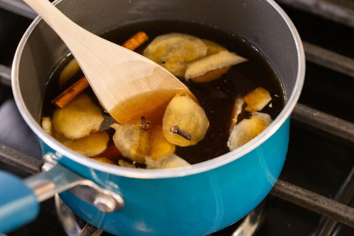 A close shot of gingerbread simple syrup simmering in a teal saucepan with sliced ginger, a cinnamon stick, and a wooden spoon.